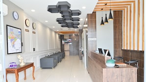 A reception area with a wooden reception desk, grey sofas, a map on the wall, clocks, and a TV screen. - Hotel Devaan (Bathinda)