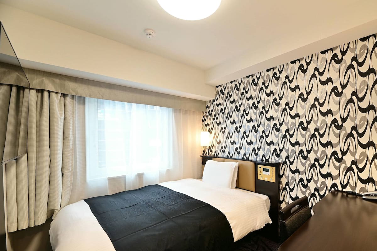 Een hotelkamer met een groot bed, een nachtkastje met lamp, een behang met patroon, en een raam met gordijnen.