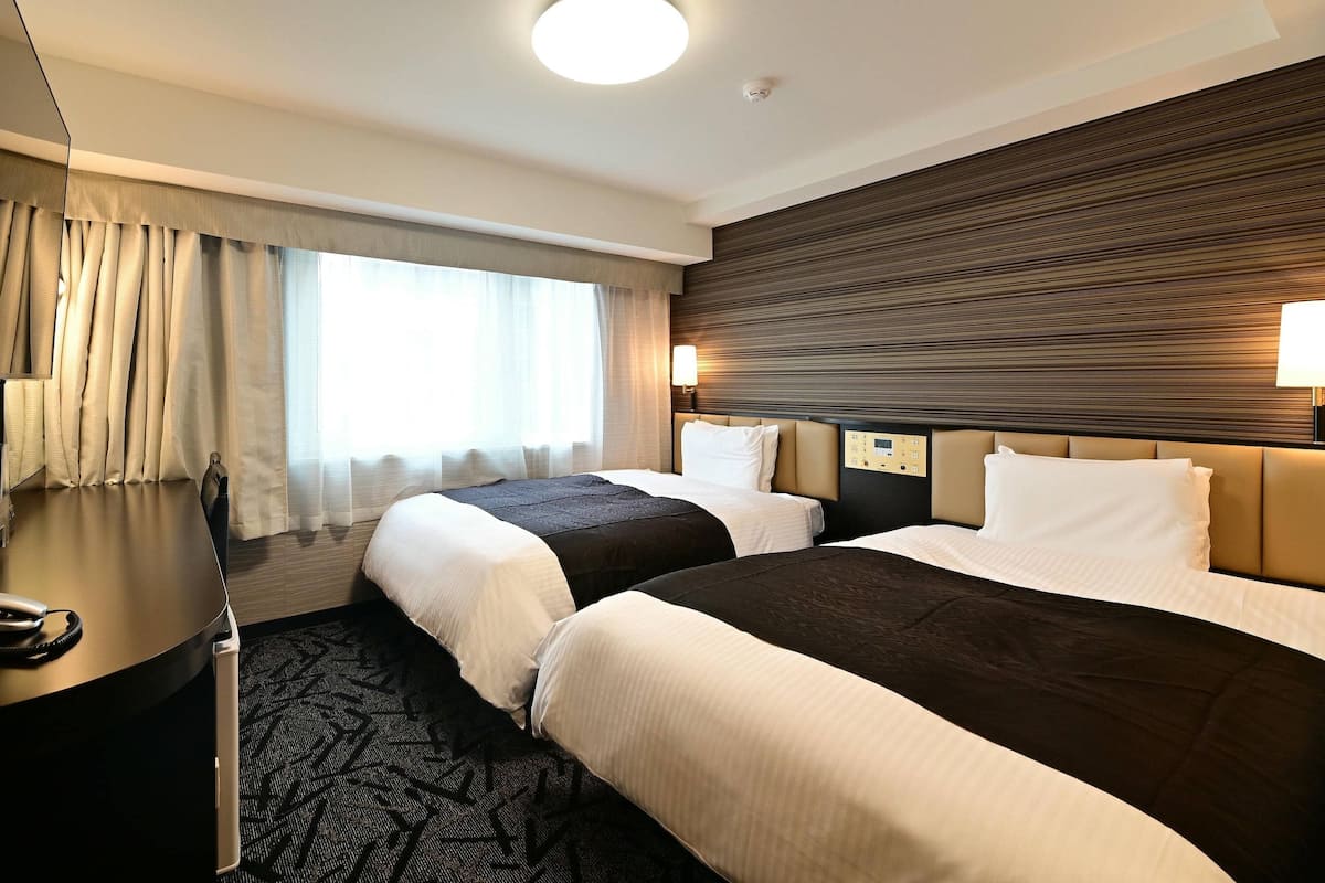Een hotelkamer met twee bedden, een bureau, een lamp en een raam met gordijnen.