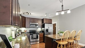 A modern kitchen with dark wood cabinets, stainless steel appliances, a granite countertop, and a central island with bar stools. - The Classic City Collection - 3 Separate Units (Athens)
