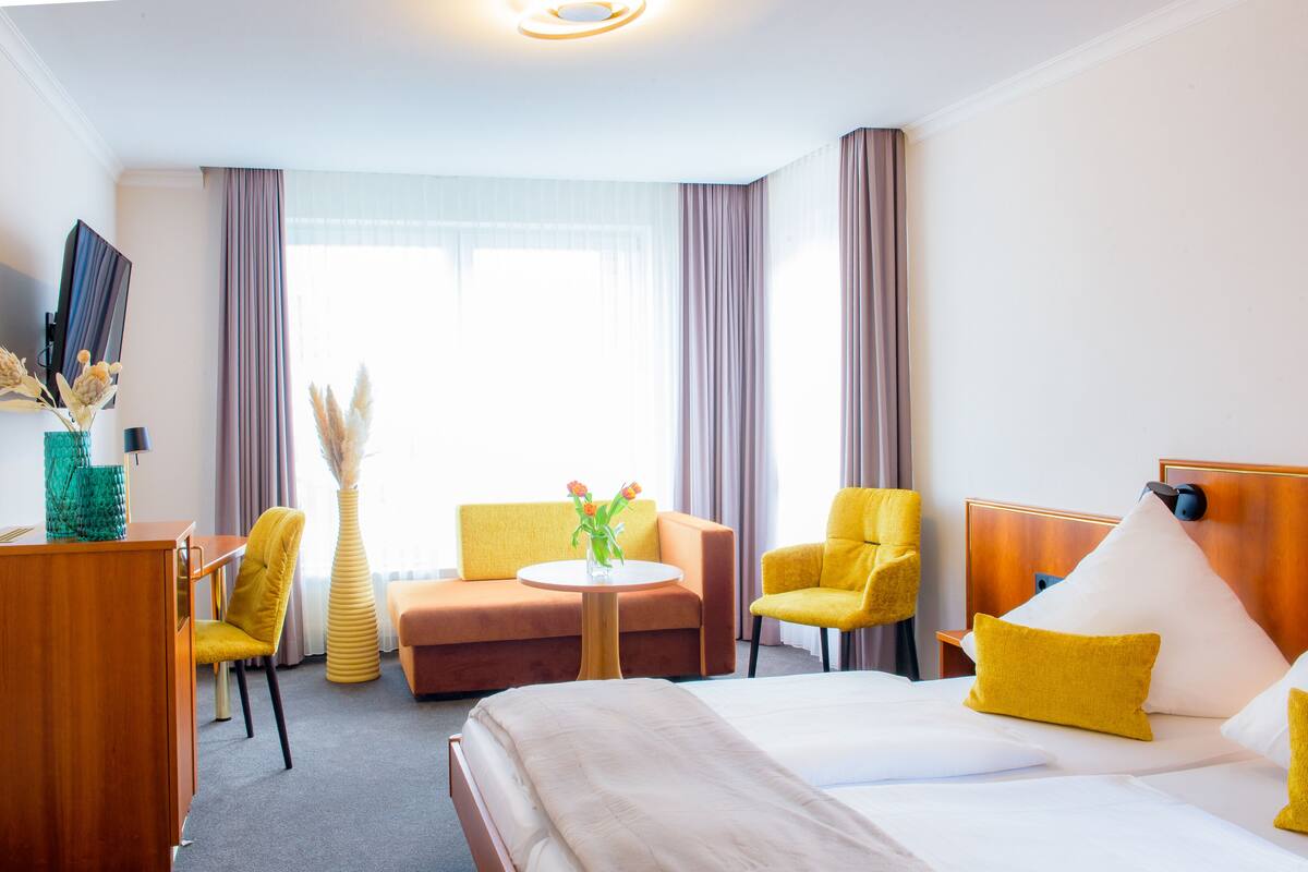 Et hotelværelse med en seng, to gule lænestole, en sofa, et lille rundt bord med en vase blomster og et fjernsyn på et træskab.