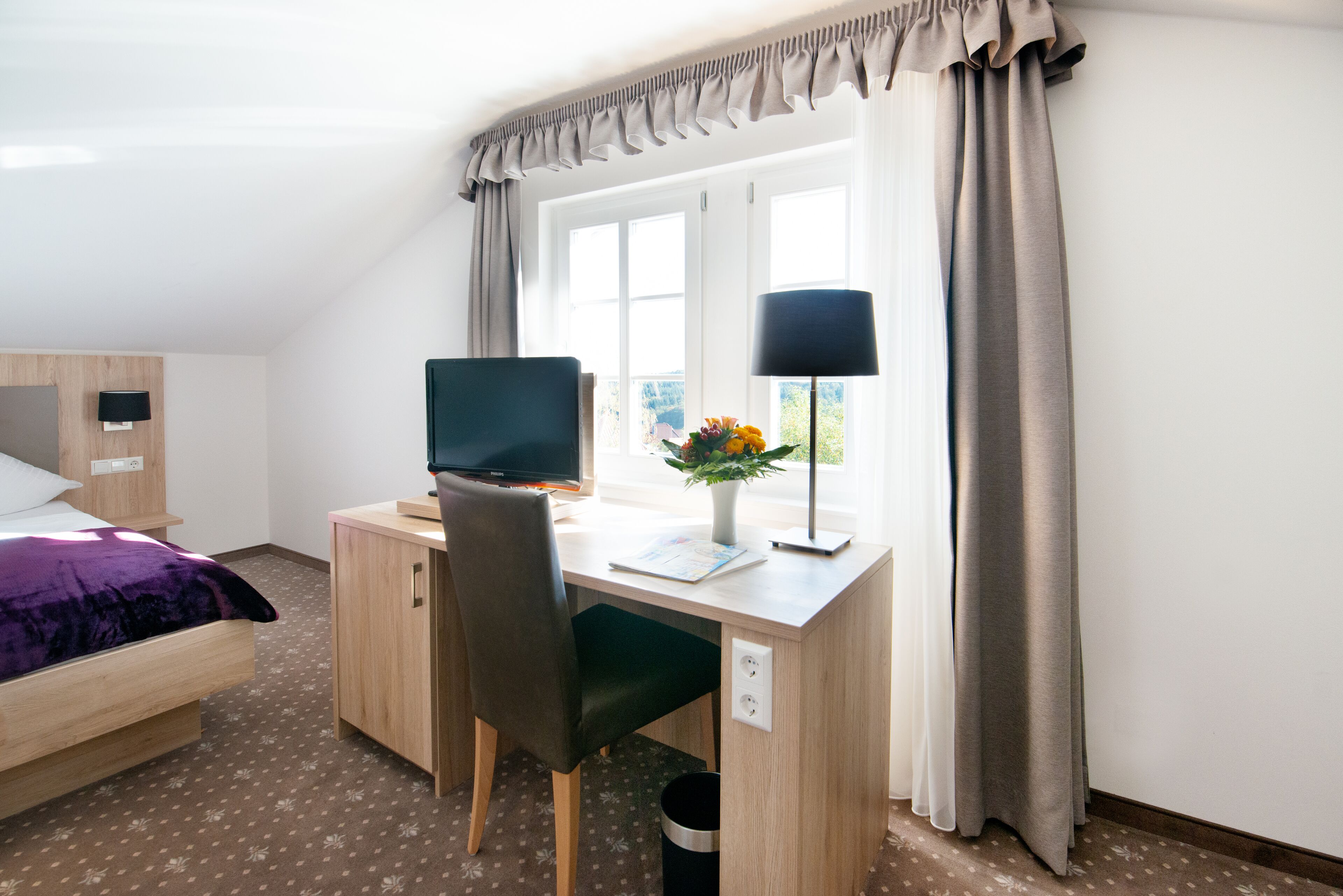 A hotel room with a bed, a desk with a computer, a chair, a lamp, and a window with curtains.