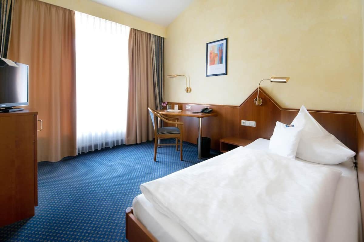 Et hotelværelse med seng, skrivebord, stol, fjernsyn og et vindue med gardiner.