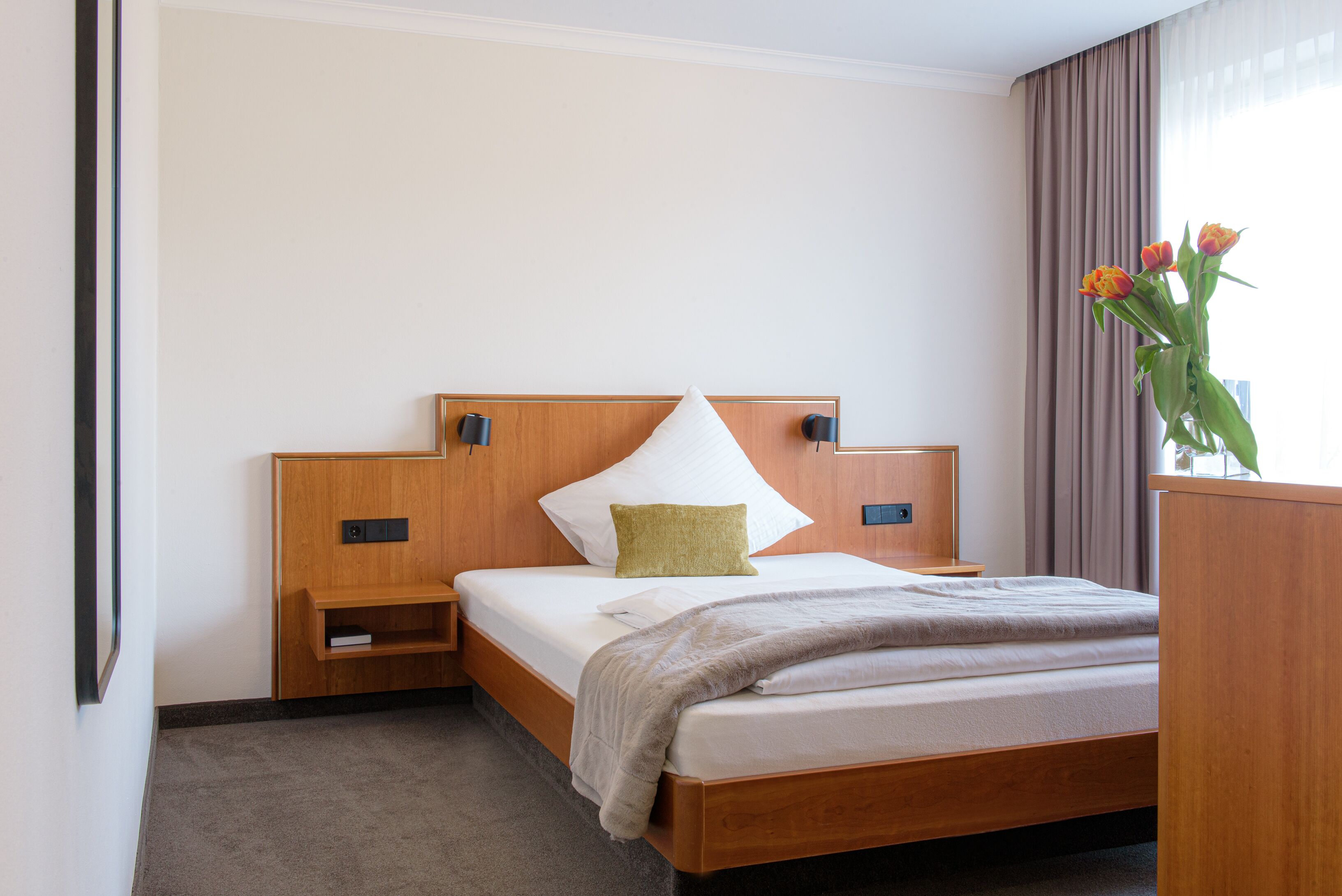 A hotel room with a wooden bed, a nightstand, a bedside table, and a vase with flowers.