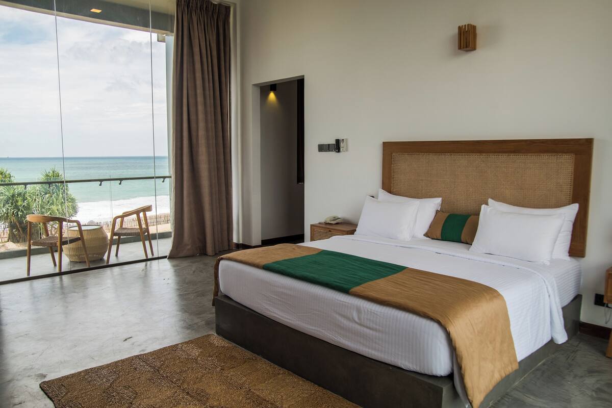 Ein Hotelzimmer mit einem großen Bett, Blick auf den Strand und einem Balkon mit Sitzgelegenheiten.