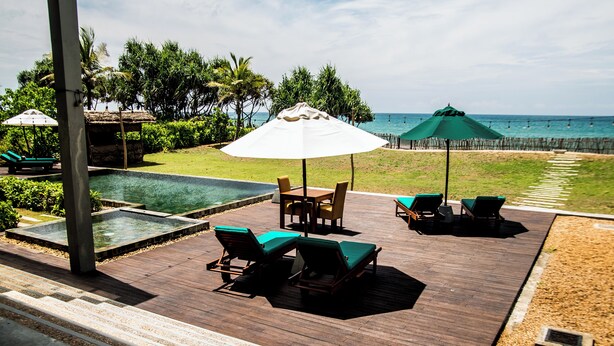 Terraza de madera con sillones de descanso y sombrillas, con vista a una piscina y al océano.