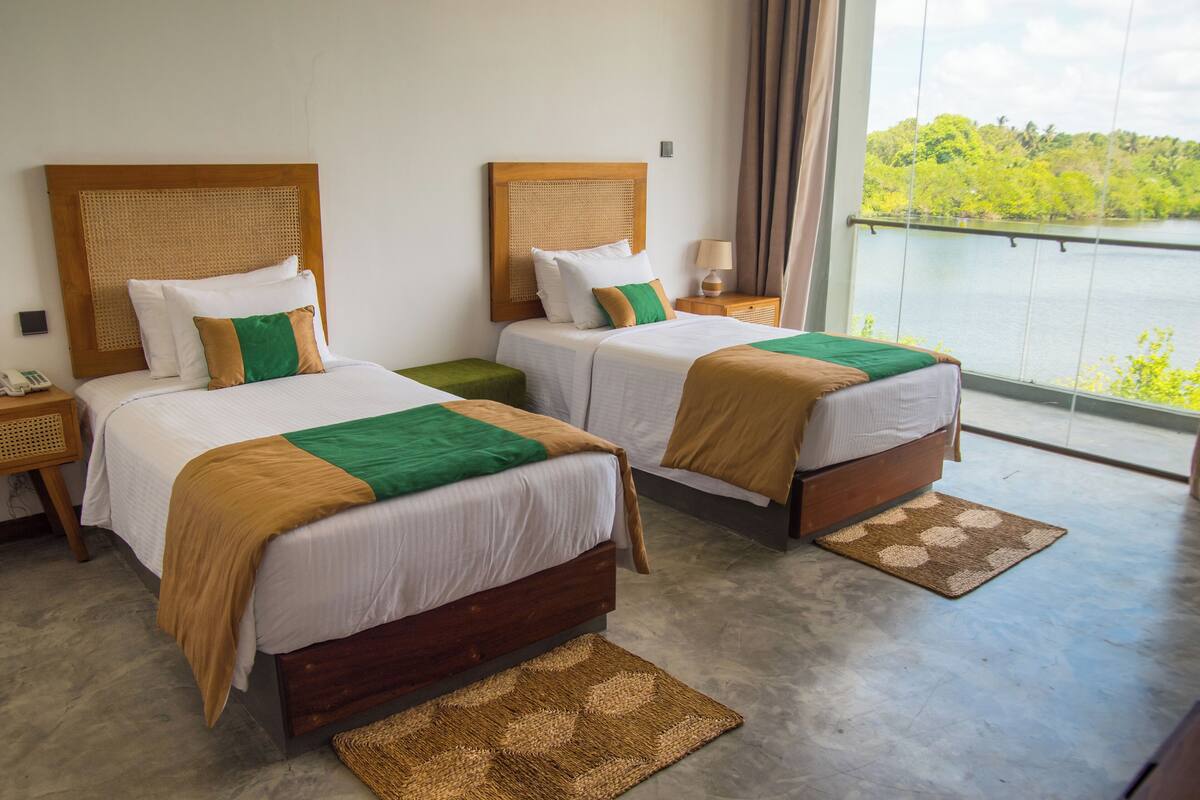 Ein Hotelzimmer mit zwei Betten, jeweils mit einem Holz-Kopfteil, weißer Bettwäsche und einem grünen Akzentkissen. Am linken Bett befindet sich ein kleiner Nachttisch mit einer Lampe und einem Telefon. Das Zimmer verfügt über ein großes Fenster mit Blick auf Grünflächen und ein Gewässer.