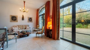 A spacious living room with a sofa, chairs, a coffee table, and a chandelier. Large windows with curtains offer a view of the outdoors. - The Hôtel de Beaumont carriages (Valognes)