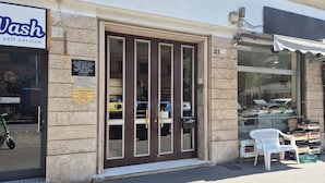 A storefront with a 'Wash self service' sign, a large glass door, and a small outdoor seating area with a white plastic chair. - Nestop Colli Albani (Rome)