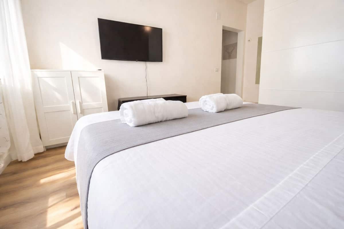Habitación de hotel con una cama grande, dos toallas enrolladas, un televisor de pantalla plana en la pared, un armario y una puerta que conduce a otra habitación.