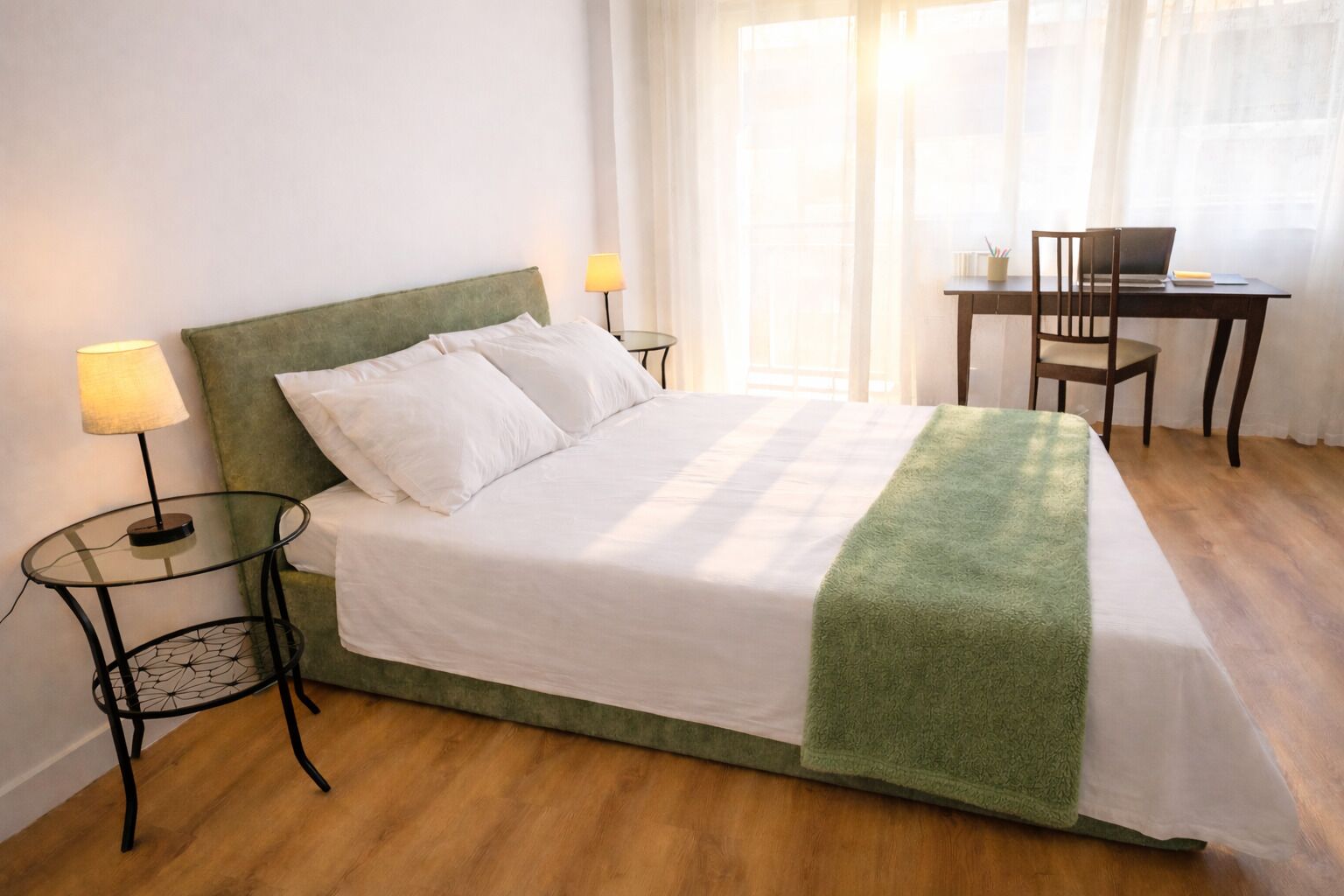 A neatly made bed with a green blanket, two bedside lamps, a small round table, and a chair with a laptop.