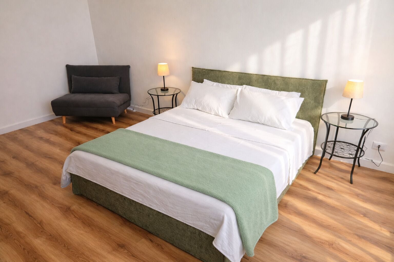 Una cama con cabecera verde, un sillón gris, dos mesitas de noche con lámparas y un suelo de madera.