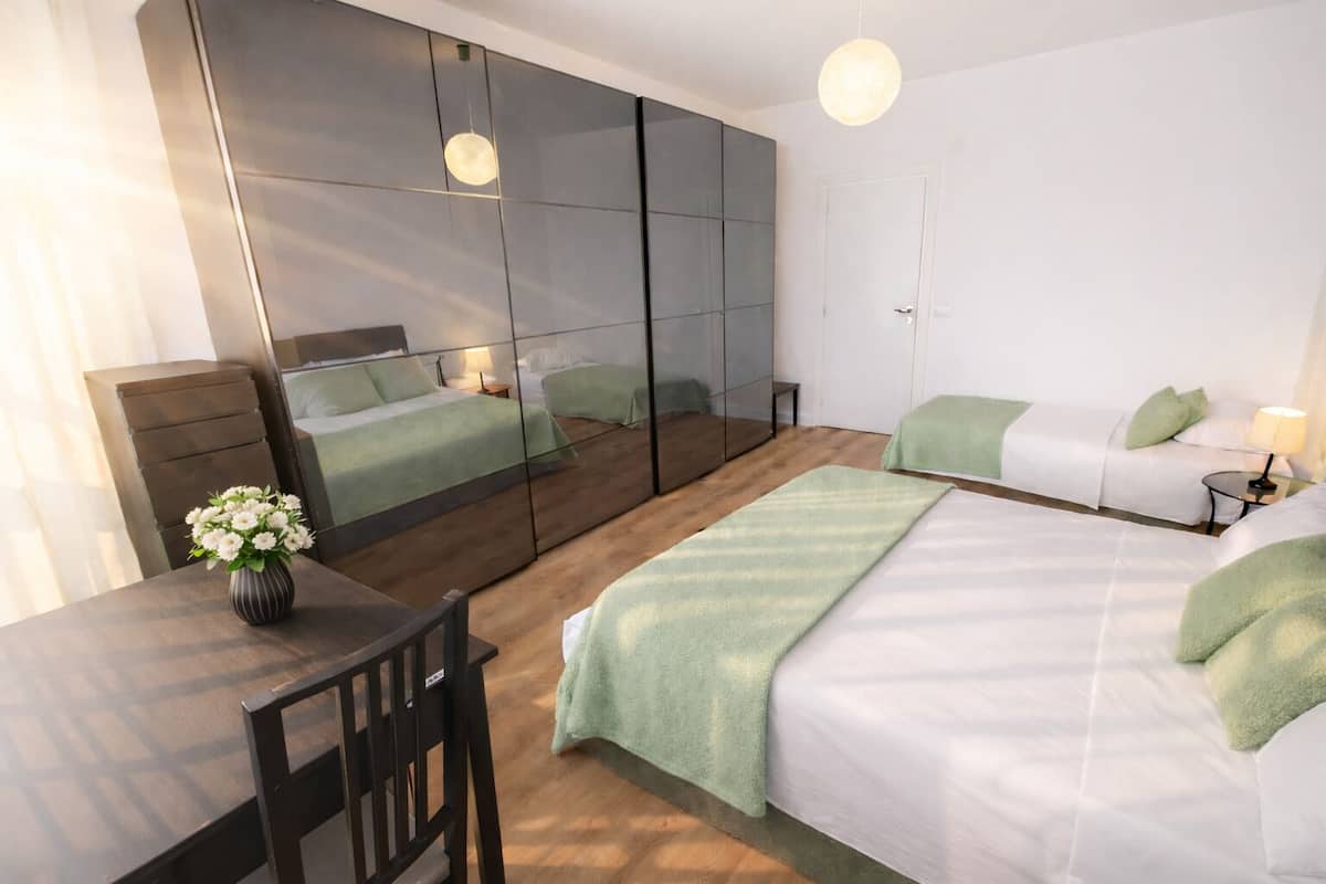 Habitación de hotel moderna con una cama grande, un escritorio y una silla. Hay un espejo en la pared, una mesita de noche con una lámpara y un jarrón con flores.