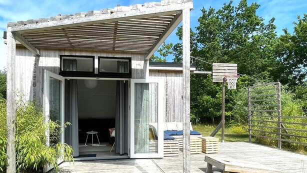 A modern wooden cabin with a sliding glass door entrance, a small patio with a bench, and a basketball hoop.