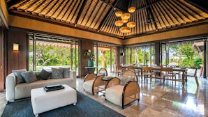 Amplia sala de estar con un sofá moderno, reposapiés y sillas de mimbre. Amplias ventanas ofrecen vistas al exterior, con abundante vegetación.