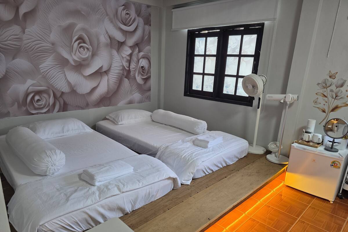 Habitació d'hotel amb dos llits, un mural floral gran a la paret, una finestra amb persianes, una taula de nit amb una làmpada i un petit armari amb articles de tocador.