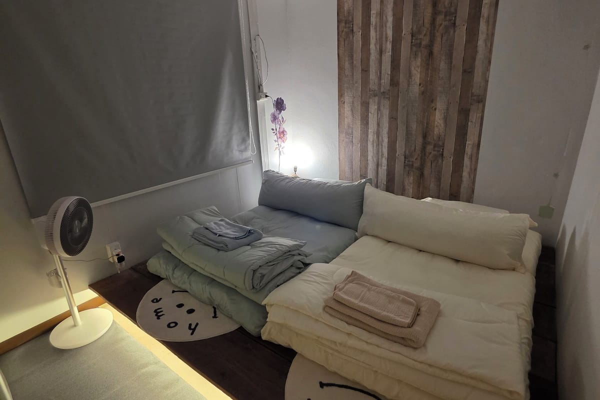 Un dormitori petit i acollidor amb un llit, una taula de nit, un ventilador i una finestra amb cortina.