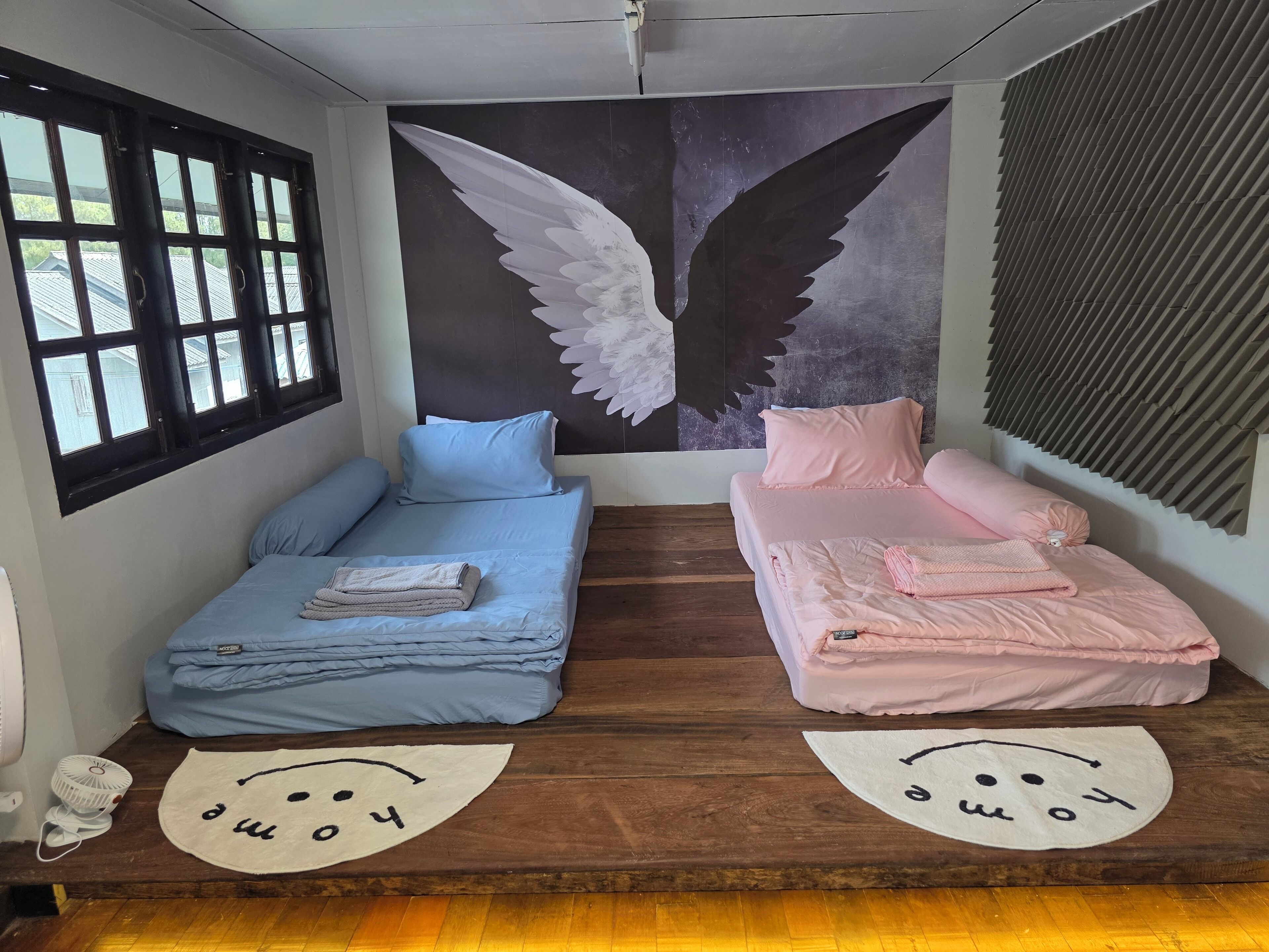 Una habitación con dos camas, una con ropa de cama azul y otra con ropa de cama rosa, un mural grande en la pared con alas y suelo de madera.