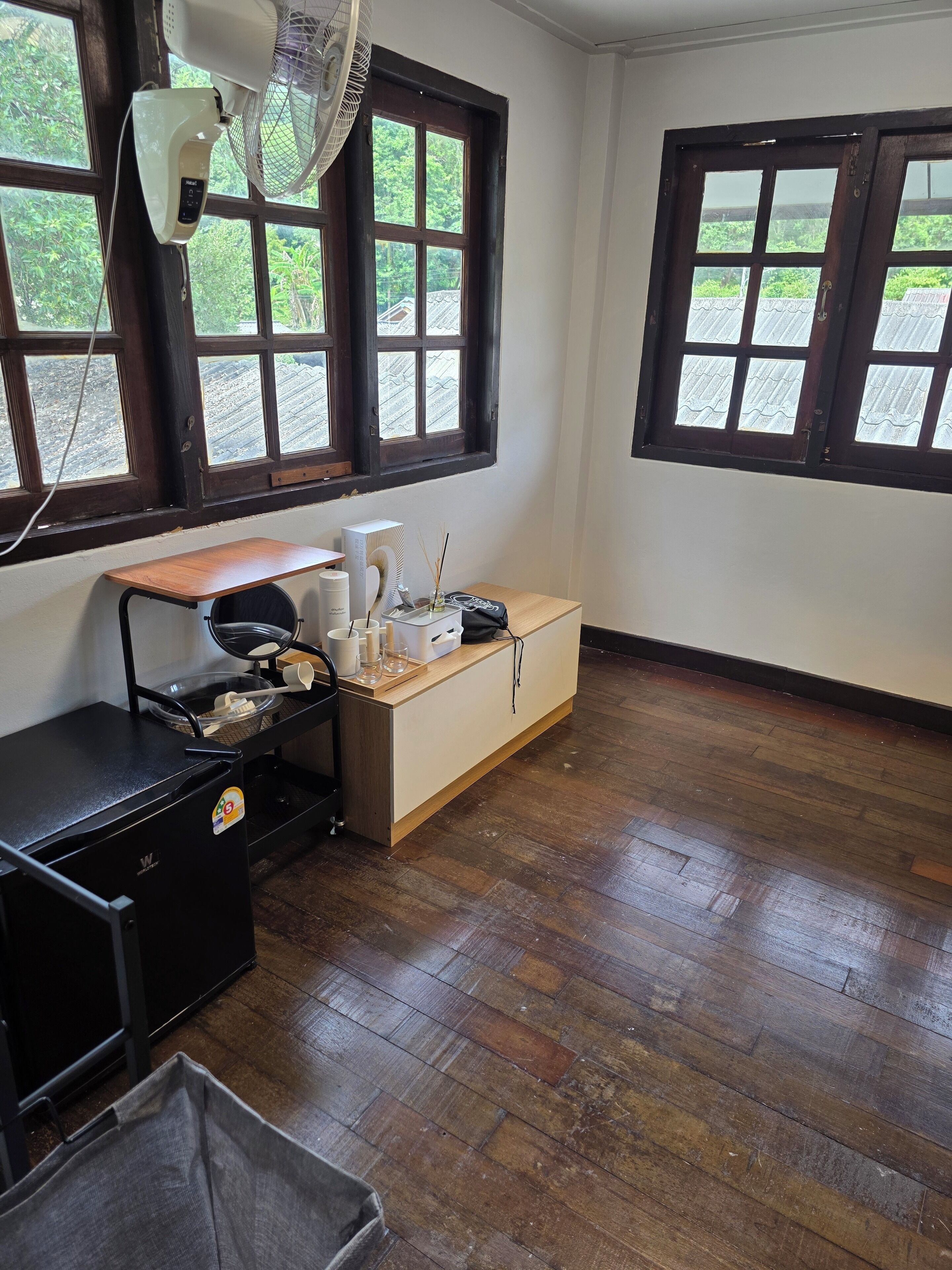 A room with a wooden floor, a desk with a computer, a fan, and a window with a view of greenery.