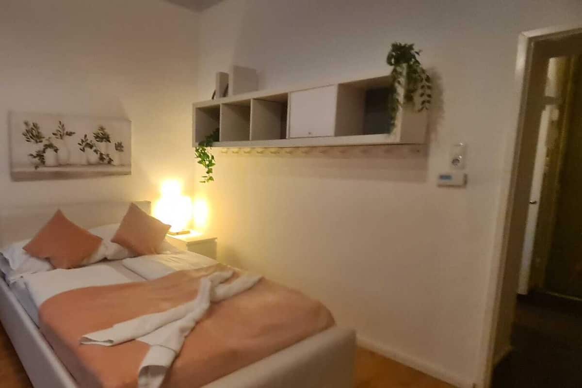 Un dormitorio con cama, mesita de noche, estante en la pared con plantas y una puerta.