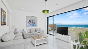 Una sala de estar moderna con un gran ventanal con vista a una playa, un sofá, una mesa de centro y un televisor.
