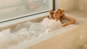 Una persona relajándose en una bañera llena de burbujas, con un amplio ventanal que ofrece vistas a la playa y al paisaje urbano.