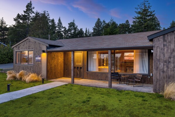 Une cabane moderne en bois, dotée d’une grande porte vitrée donnant sur un coin repas, le tout entouré d’une pelouse bien entretenue.