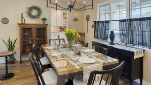 A dining area with a long table set for six, surrounded by chairs. There is a chandelier above the table, a window with patterned curtains, and a cabinet with glass doors to the left.