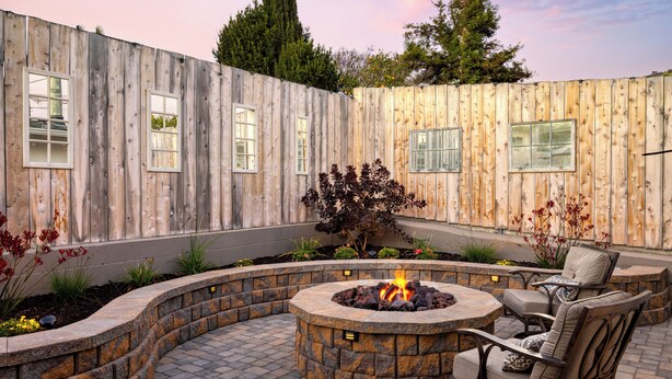 A circular stone fire pit with a fire, surrounded by a stone wall and a wooden fence. There are two chairs and a table near the fire pit.