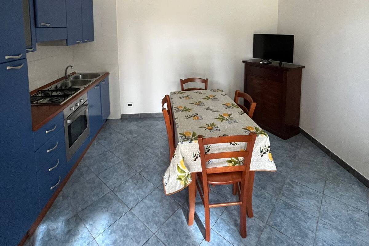 Eine kleine Küche mit blauen Schränken, einem Esstisch mit Stühlen, einem Fernseher auf einem Schrank und einem Fliesenboden.