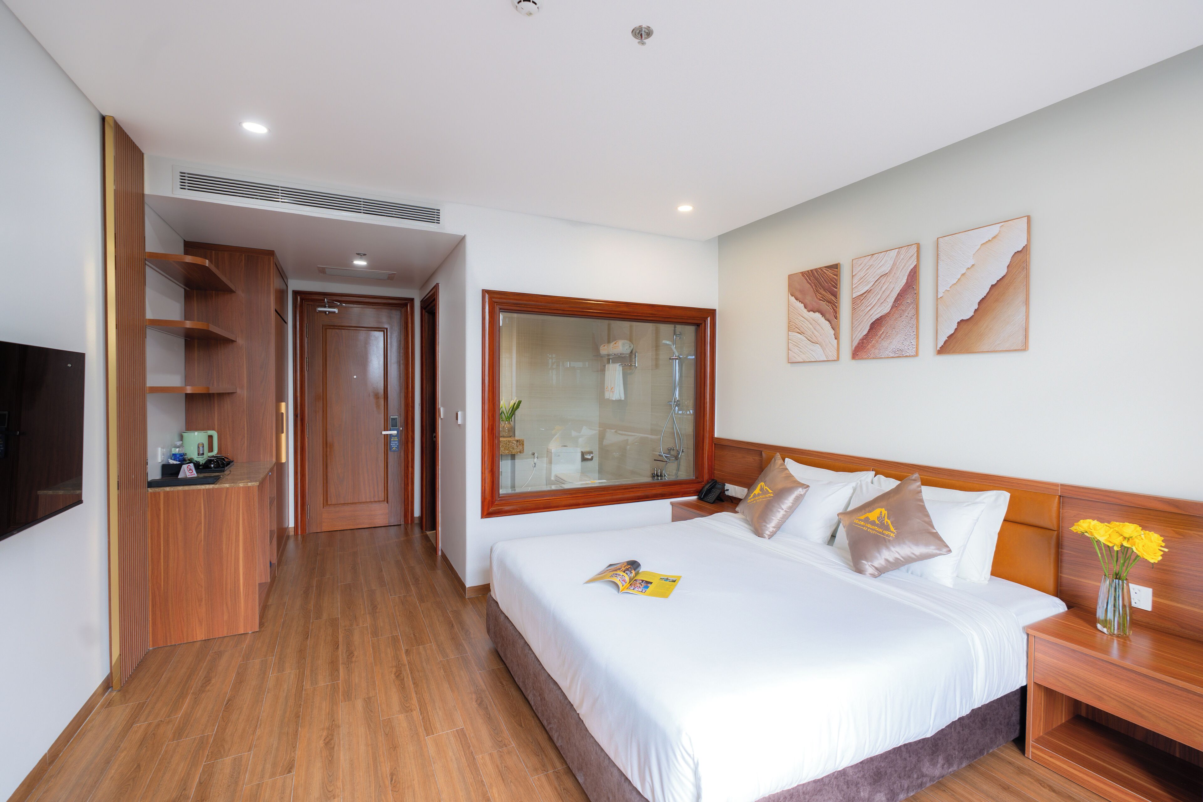 A hotel room with a large bed, wooden headboard, a bedside table with a vase of flowers, a large window, and a built-in TV.