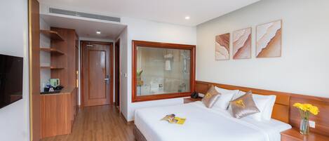 A hotel room with a large bed, wooden headboard, a bedside table with a vase of flowers, a large window, and a built-in TV.