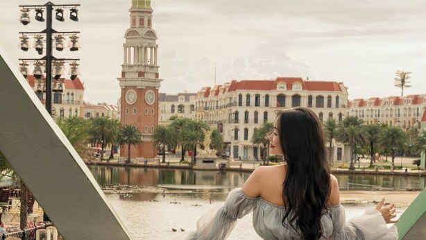 Uma pessoa está em uma ponte, com vista para um edifício histórico com uma torre do relógio.
