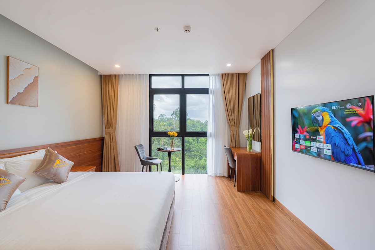 Une chambre d’hôtel équipée d’un lit, d’un bureau, d’une chaise, d’une télévision et d’une fenêtre donnant sur un paysage verdoyant.