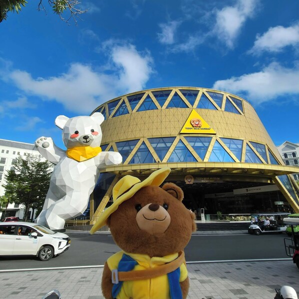 A teddy bear mascot in front of a building with a yellow triangular logo and the text "TEDDY BEAR MUSEUM". - Golden Mountain Hotel Phu Quoc (Phu Quoc)