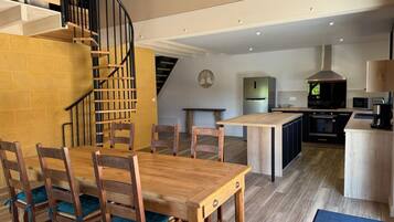 Eine moderne Küche mit Kochinsel, einem Essbereich mit Holzsesseln und einem Tisch sowie einer Wendeltreppe, die in eine obere Etage führt.
