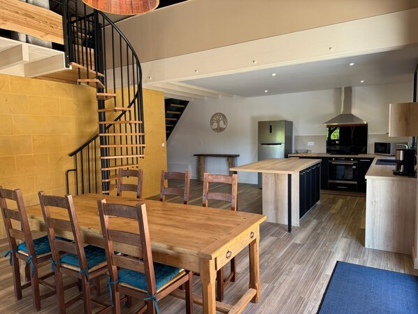 Una cocina moderna con una isla central, un comedor con sillas de madera y una mesa, y una escalera en espiral que conduce a un nivel superior.