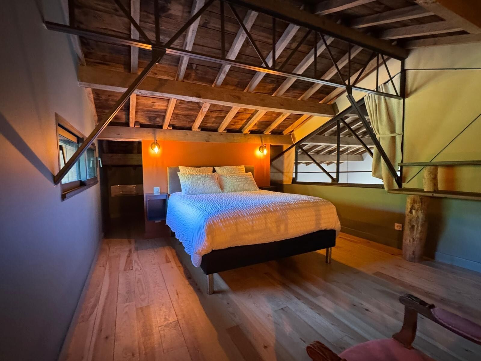 Un dormitorio con techo inclinado, vigas de madera, una cama con cubrecama blanco y una mesita de noche pequeña.