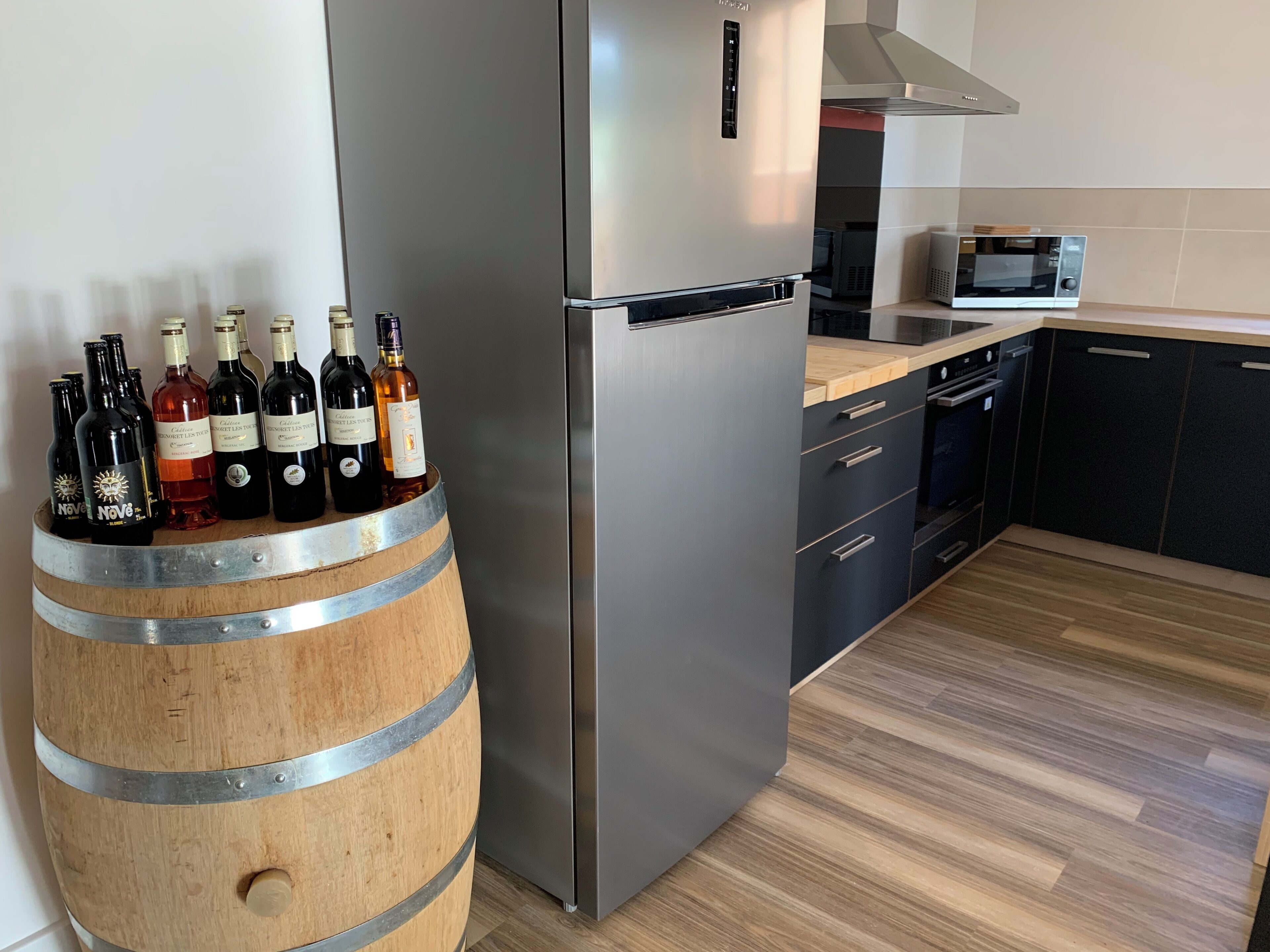 A modern kitchen with a stainless steel refrigerator, a wooden barrel with wine bottles, and a microwave.
