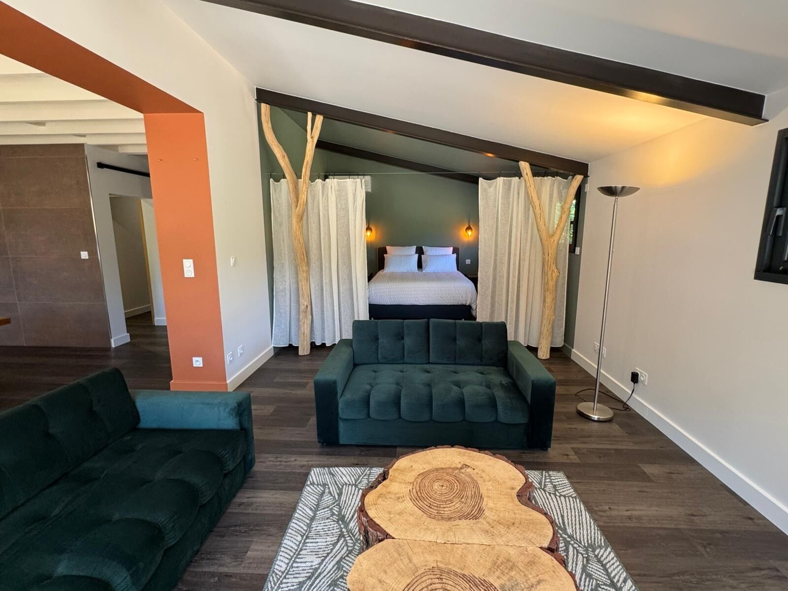 Un salon moderne avec un canapé, une table basse et une vue sur une chambre à coucher, où l’on aperçoit un lit et des rideaux.