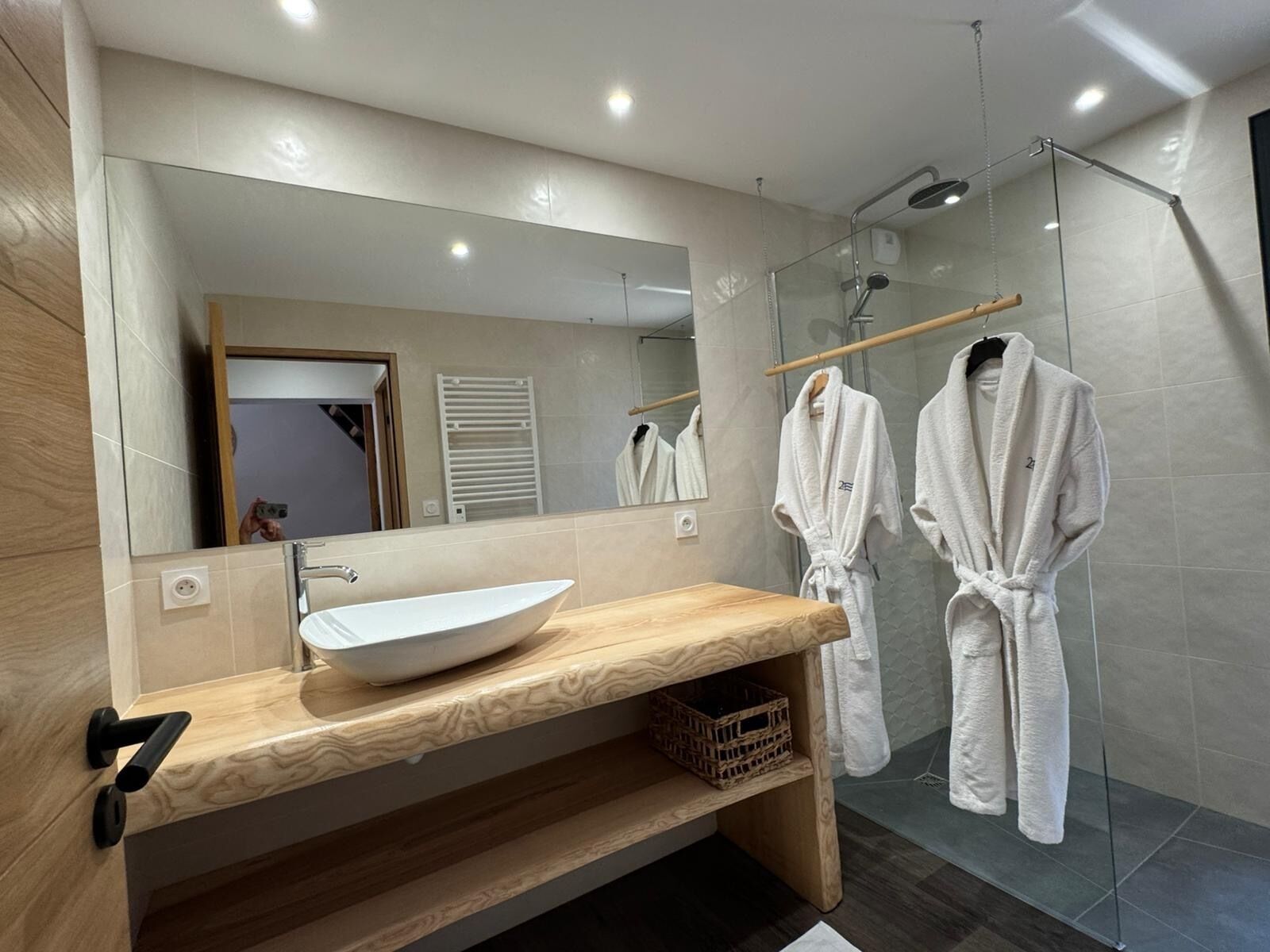 Un baño moderno con un espejo grande, un lavabo sobre encimera, tocador de madera, dos albornoces blancos y una ducha con mampara de cristal.