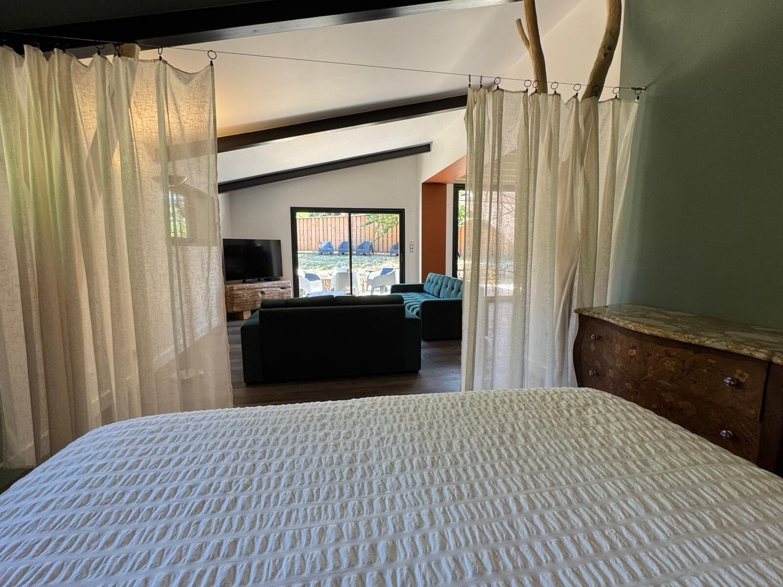 A bedroom with a large bed, a wooden dresser, and a TV in the background.