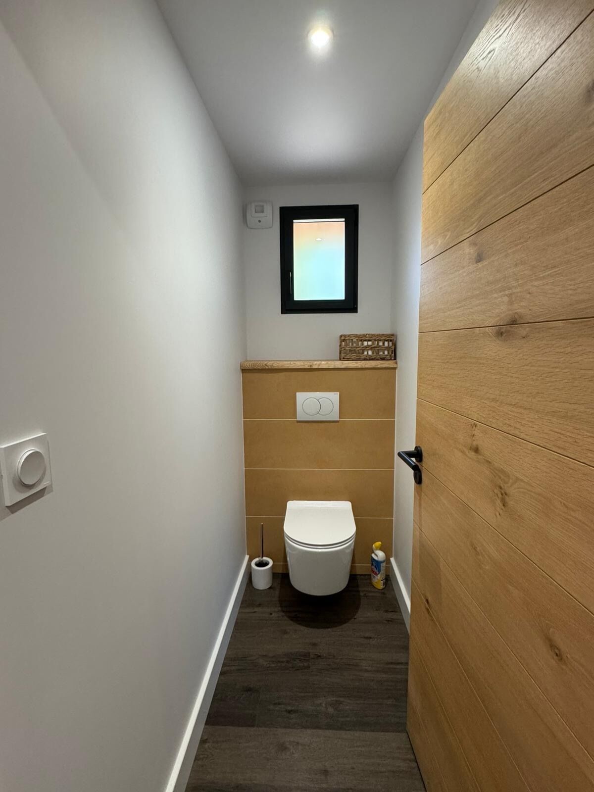 A narrow bathroom with a toilet, a small window, and a wooden door.