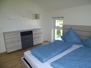 Ein Schlafzimmer mit Bett, Kommode, Fenster und Blick ins Freie.
