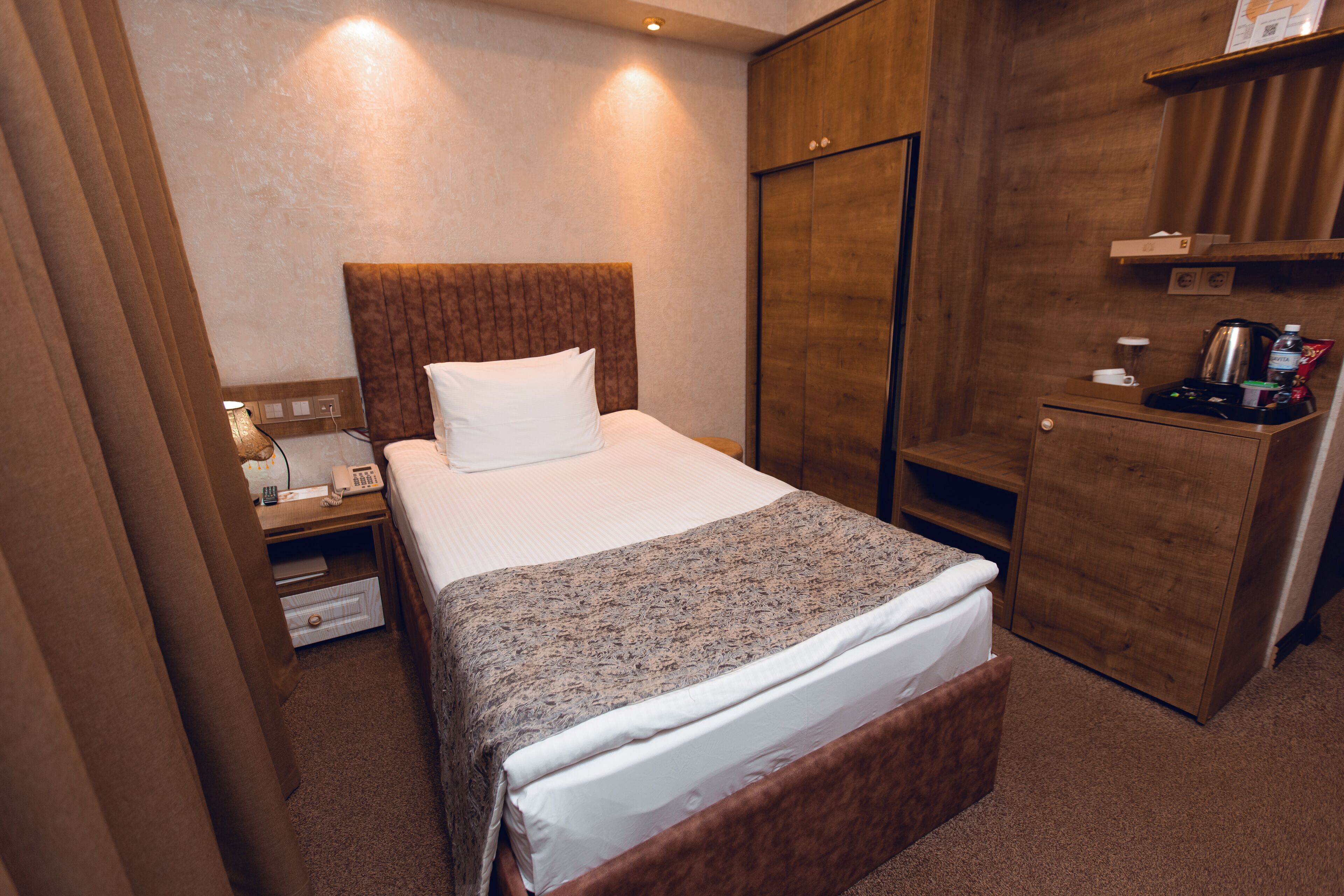 A hotel room with a bed, a wooden wardrobe, a nightstand with a lamp, and a small table with a coffee maker.