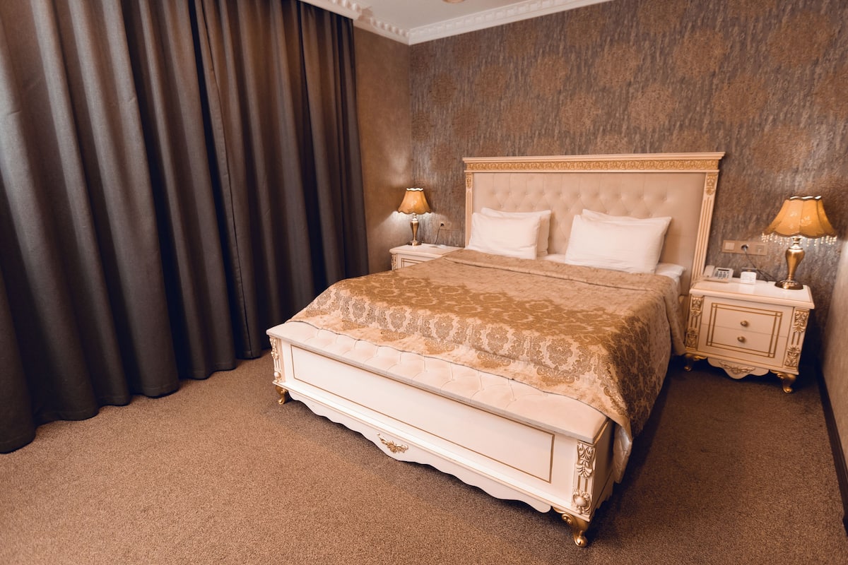 Un grand lit avec une tête de lit capitonnée, deux tables de chevet avec des lampes et un rideau foncé.