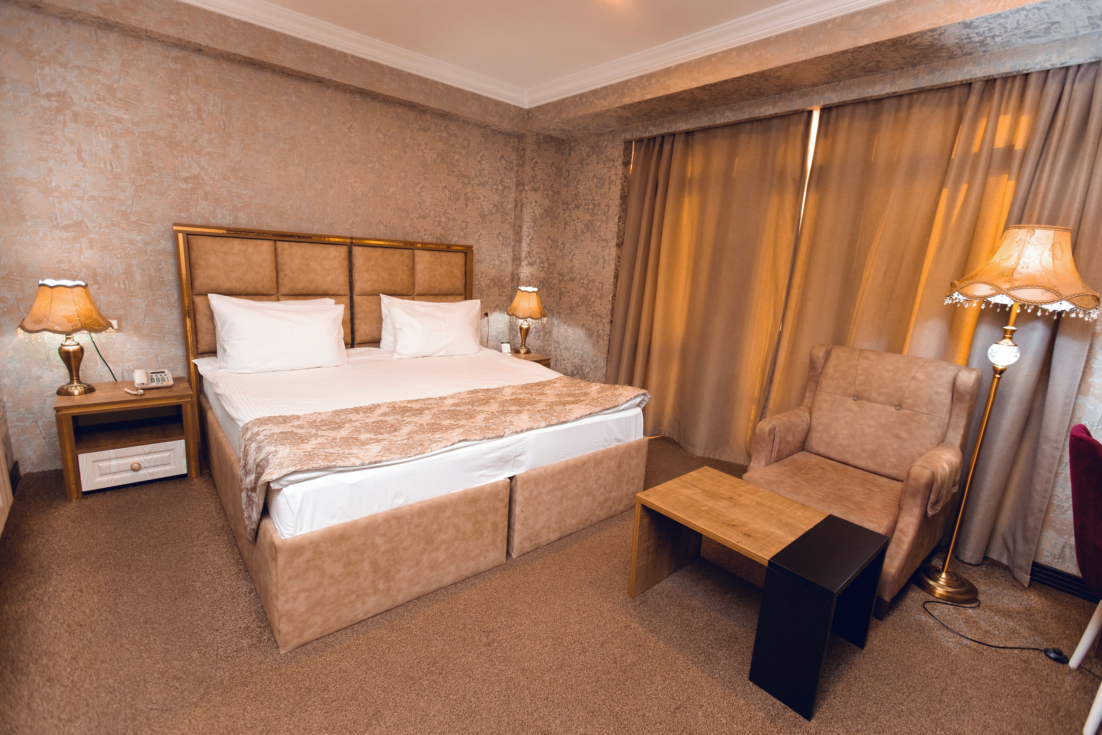 Hotellihuone, jossa on suuri sänky, kaksi yöpöytää valaisimineen, puinen sivupöytä, tuoli ja verho.