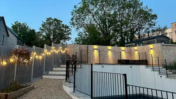 A gravel pathway leading to a wooden fence with string lights, a metal gate, and a green lawn area. - Luxury 5 Bed - Sleeps 14 - With Swimming Pool (Bristol)