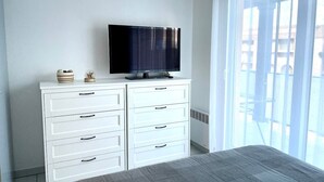Un dormitorio con cama, cómoda blanca con cajones, televisor de pantalla plana y ventana grande con persianas.