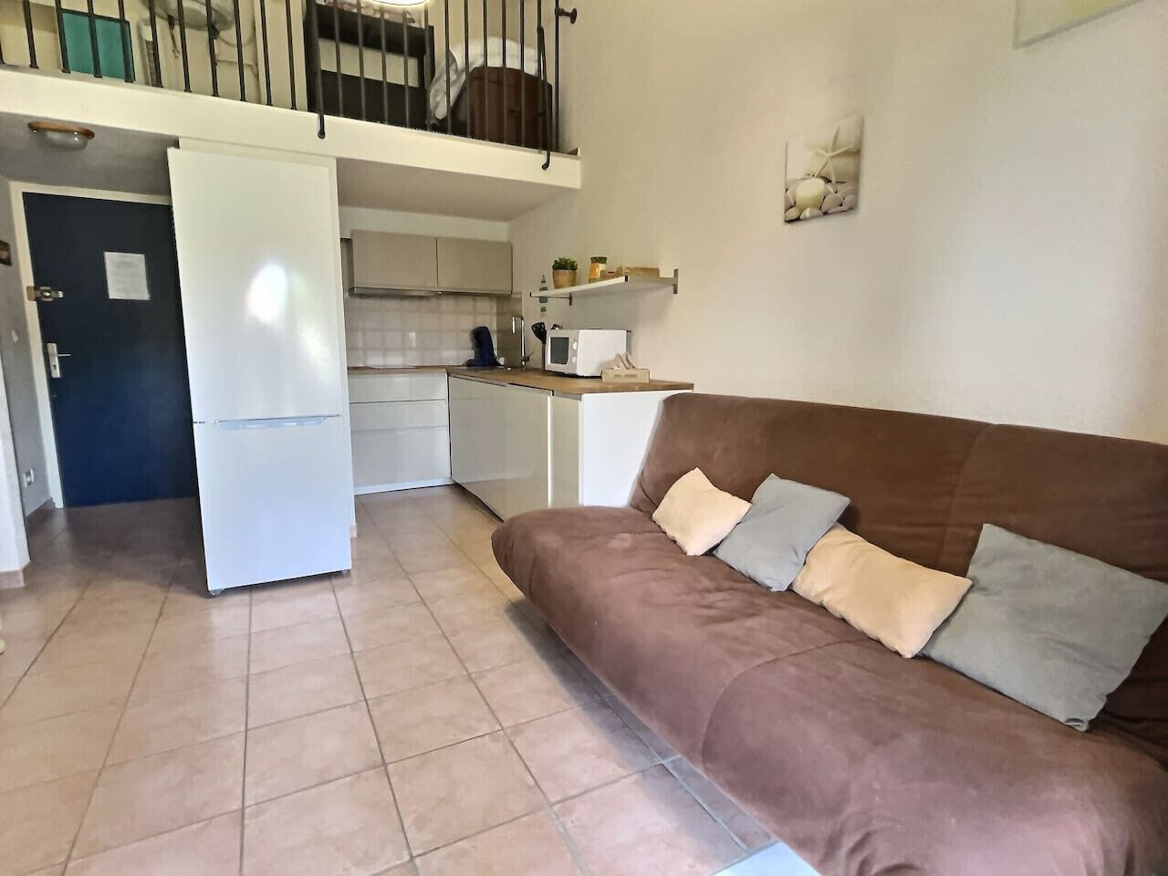 Un espacio habitable pequeño con kitchenette, un sofá marrón con cojines y una puerta que da a un nivel superior.